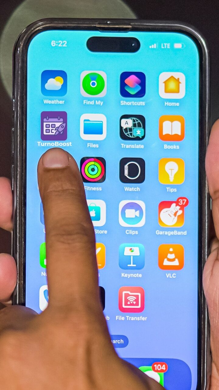 Mano sosteniendo un iPhone, dedo a punto de tocar el ícono de TurnoBoost en la pantalla de inicio, junto a Weather, Find My, Shortcuts y otras apps del sistema.